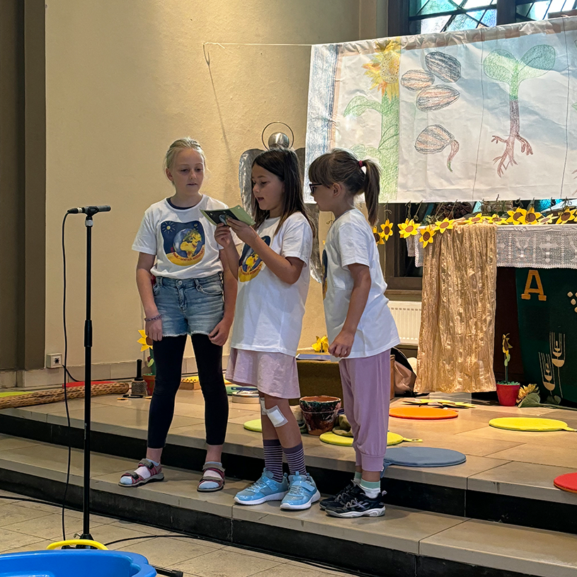 Drei Kinder aus der OGS Wasserfuhr stehen ganz vorn in der festlich, farbenfrohen geschmückten Kirche und lesen etwas vor.